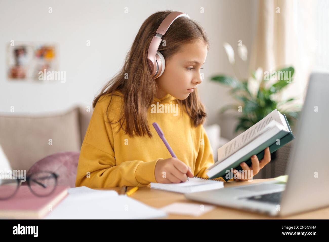 Scuola ragazza studiare online. Scuola a casa. Educazione a distanza. La bambina sta leggendo il libro, facendo i compiti a casa, usando il laptop e le cuffie per imparare a distanza, avendo lezione in linea con l'insegnante della scuola Foto Stock