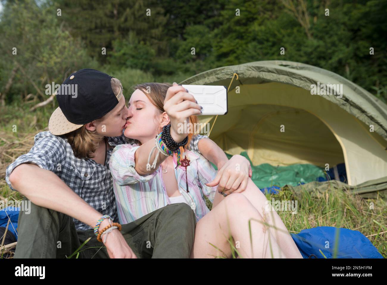 Giovane coppia baciare e prendere selfie con smartphone di fronte al camping tenda, Baviera, Germania Foto Stock