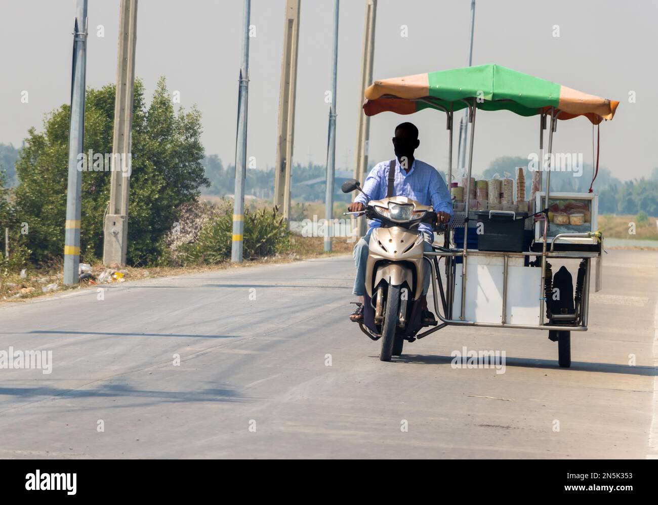 Un distributore di gelati guida una moto a tre ruote lungo una strada del villaggio, Thailandia Foto Stock