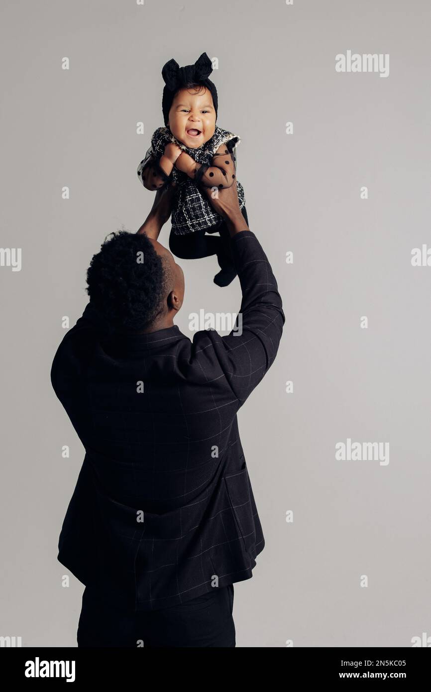 Il giovane uomo africano gioca con la figlia del bambino dal matrimonio interracial contro lo sfondo grigio. Il bambino ride allegro. Concetto di famiglia interracial Foto Stock