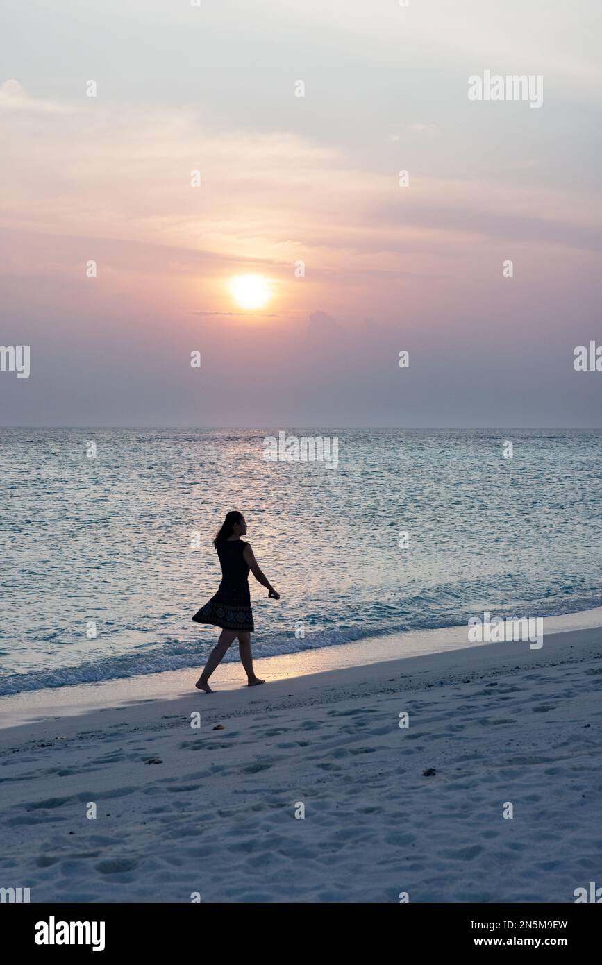 Una donna che cammina da sola su una spiaggia di sabbia al tramonto sul bordo delle acque, in silhouette, vista posteriore, le Maldive, esempio di viaggio solista stile di vita. Foto Stock
