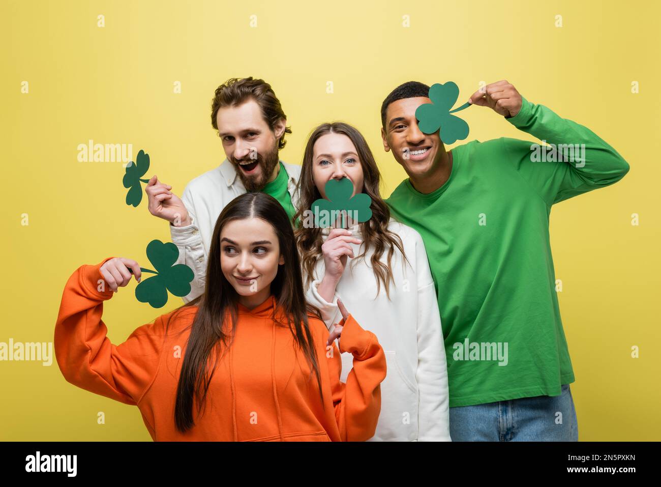 Amici multietnici che detengono trifoglio di carta durante il giorno di san patrizio isolato sul giallo Foto Stock