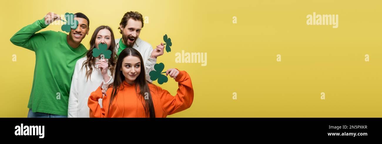 Amici interrazziali positivi che tengono verde trifoglio di carta isolato su giallo, banner Foto Stock
