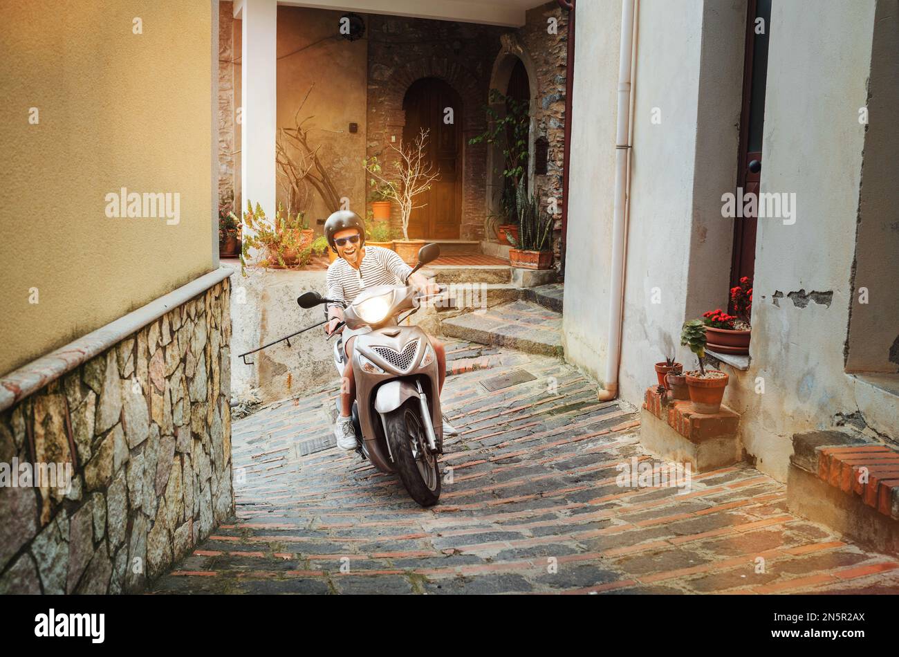 Uomo allegro e sorridente in casco e occhiali da sole che guida veloce il  moto scooter sulle strade della città vecchia siciliana nella forza d'Agro.  Felice vacanza italiana Foto stock - Alamy