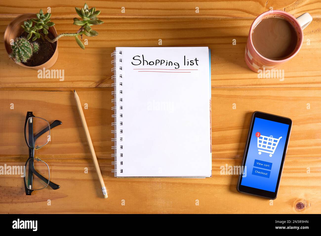 Testo della lista degli acquisti su notebook e telefono cellulare vuoti che mostra l'interfaccia dell'app del negozio online con l'icona del carrello e il pulsante di acquisto sul tavolo in legno w Foto Stock