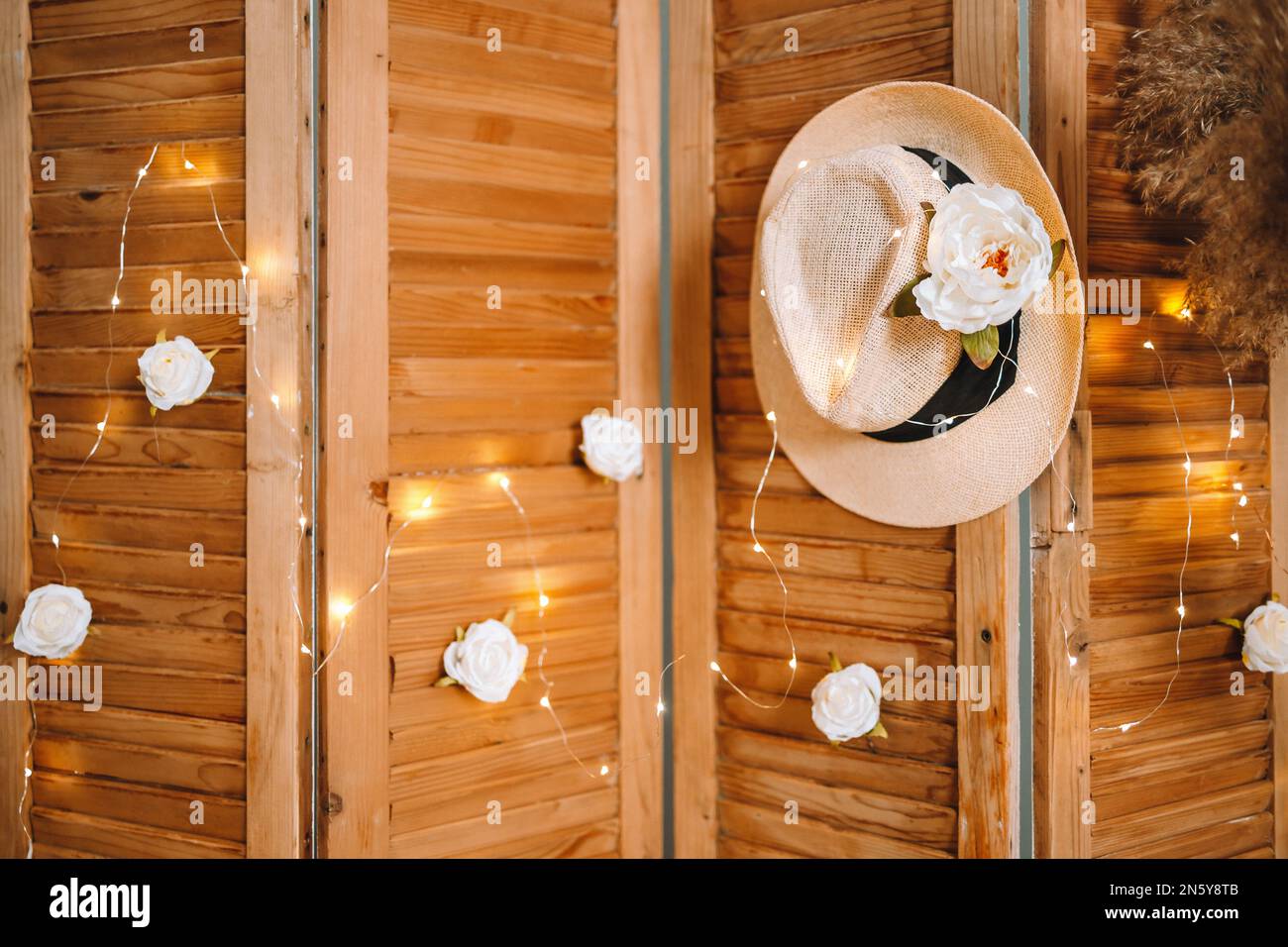 Cappello beige su uno schermo naturale pieghevole con rose bianche, pampas e calde luci a LED, accogliente e morbido Foto Stock