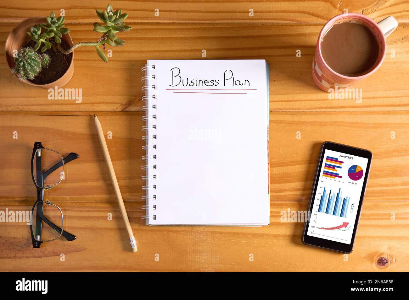 Vista dall'alto del testo del business plan sulla pagina vuota del notebook e sul telefono cellulare che mostra i grafici dei rapporti sulle prestazioni su un tavolo di legno con matita, occhiali, su Foto Stock