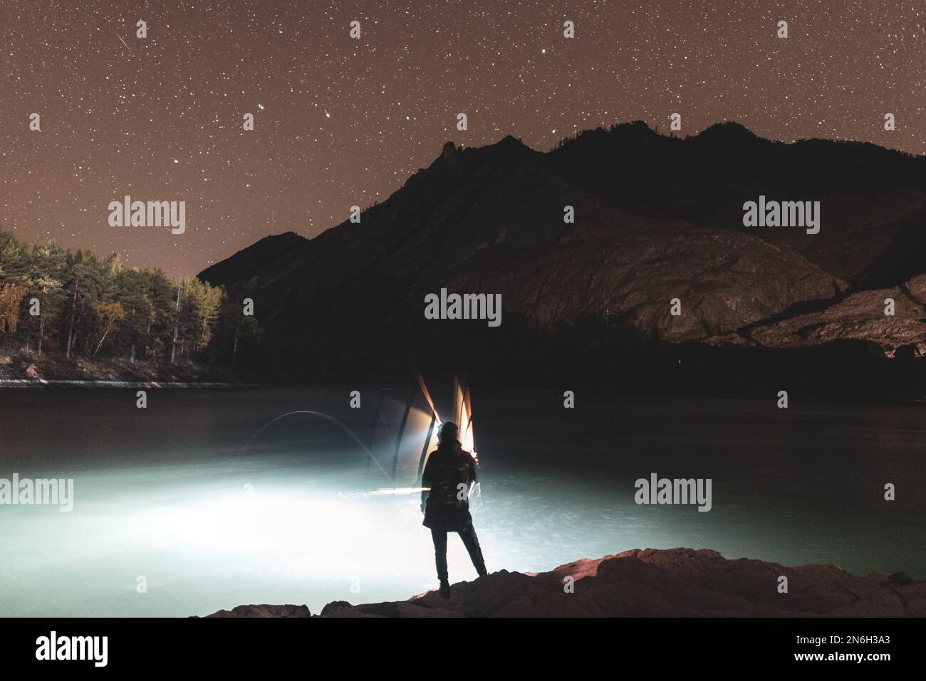La silhouette di una ragazza pescatore con una lanterna ondeggia una canna da pesca di notte sulla riva del fiume sotto una roccia con stelle in Altai in Siberia. Foto Stock