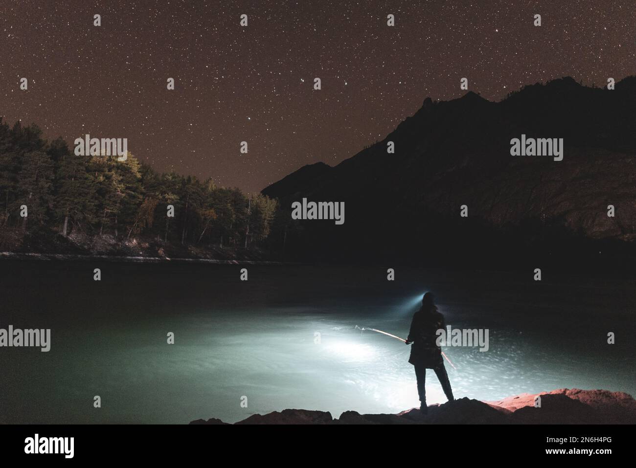 La silhouette di una ragazza pescatore con lanterna di notte si erge con una canna da pesca sulla riva del fiume sotto una roccia con stelle nell'Altai in Siberia Foto Stock