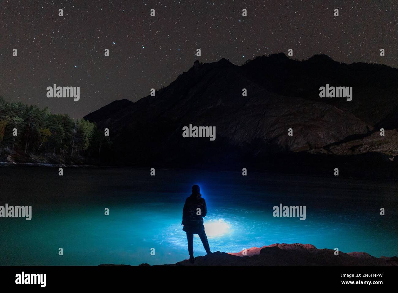 La silhouette di una ragazza pescatrice con una lanterna di notte al buio si erge con una canna da pesca sulla riva del fiume sotto una roccia con stelle ad Altai Foto Stock