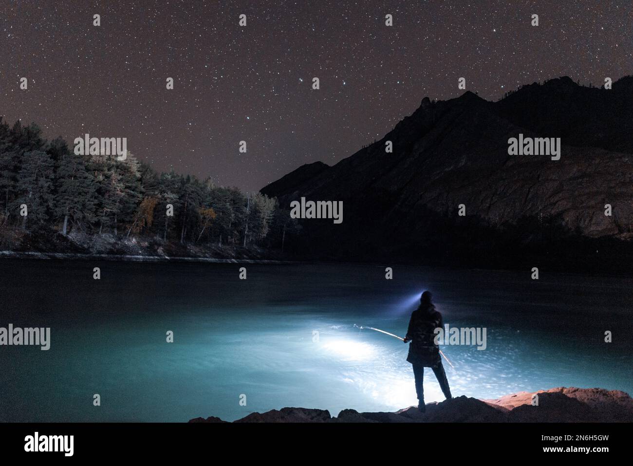 Silhouette di una ragazza pescatore con la luce di una lanterna di notte con una canna da pesca sulla riva del fiume sotto una roccia con stelle in Altai in Siberia. Foto Stock