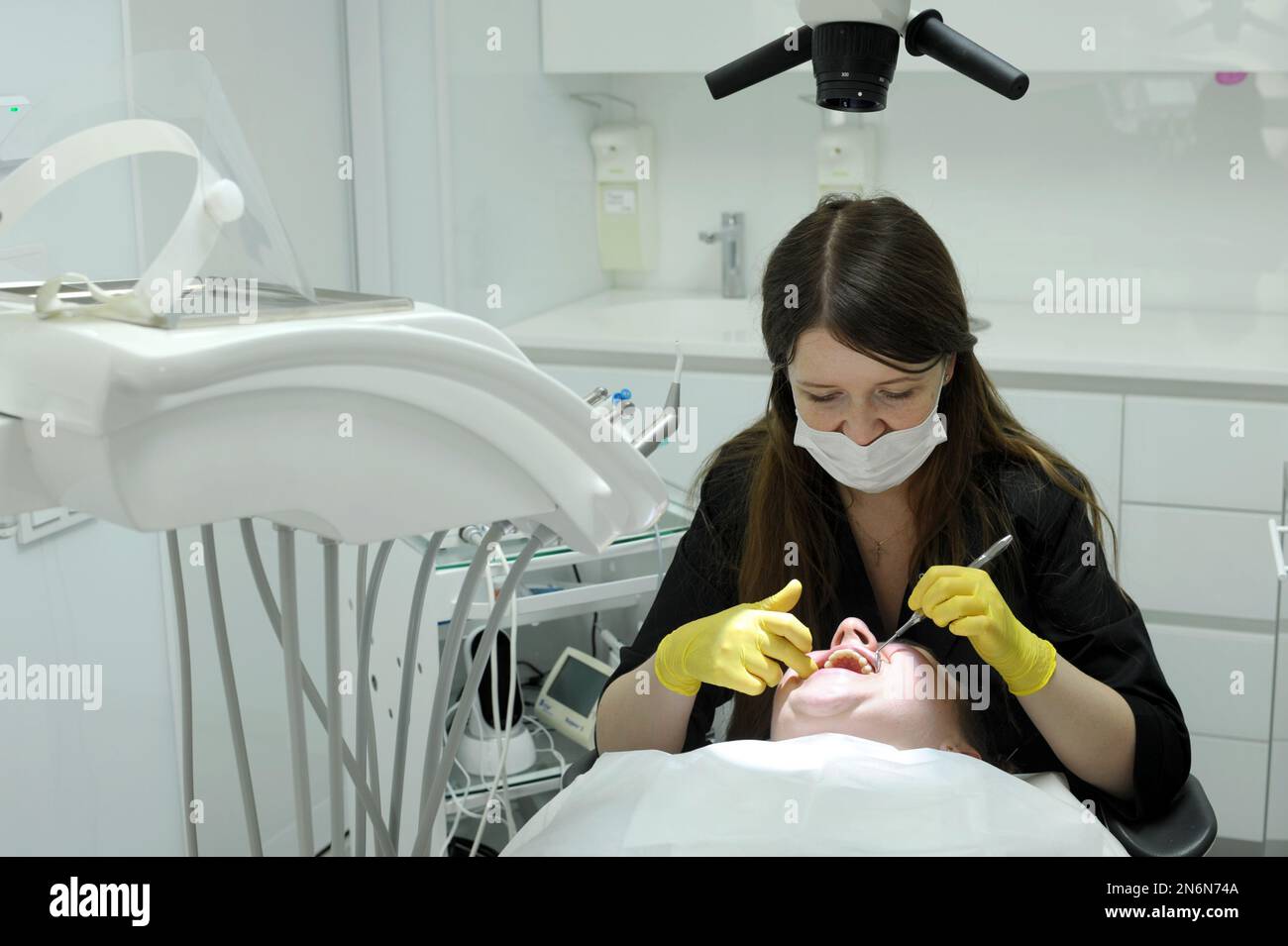 donna dentista in guanti gialli e vestiti neri denti protesici esamina il paziente con l'aiuto di specchio un sacco di ultima tecnologia whit Foto Stock