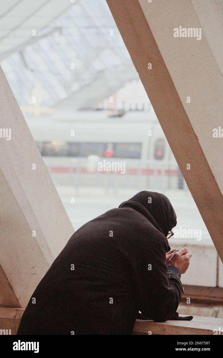 Una vista posteriore di una persona che si appoggia sul davanzale e guarda il suo telefono nella stazione ferroviaria Foto Stock