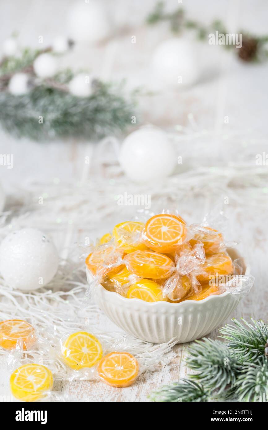 Ciotola con caramelle a fettine di arancia e limone, confezionate singolarmente in foglio, su fondo bianco con decorazione natalizia, verticale Foto Stock