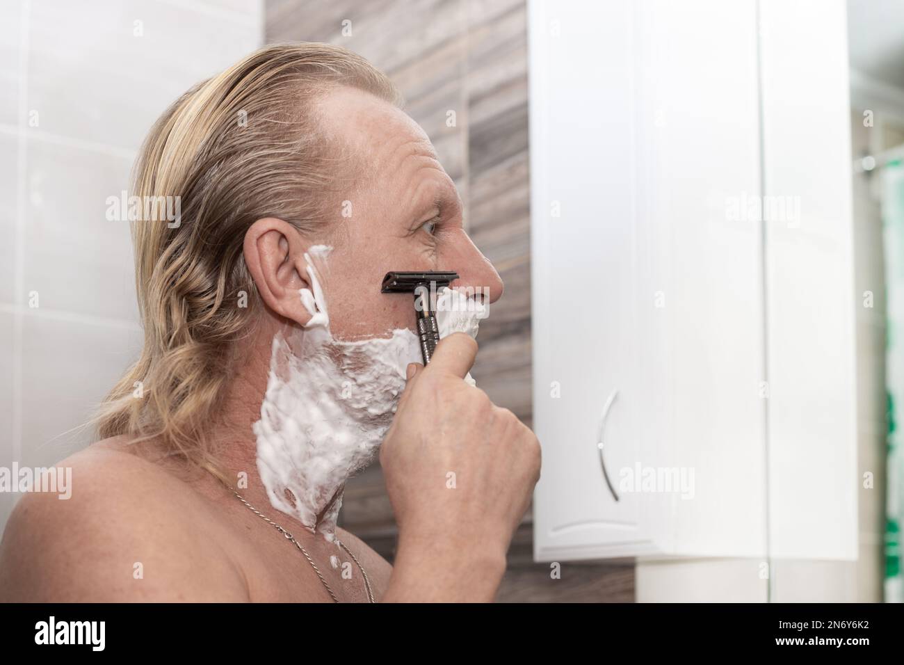 Un uomo maturo rasa con un rasoio di sicurezza in bagno. Procedure di igiene del mattino. Foto Stock