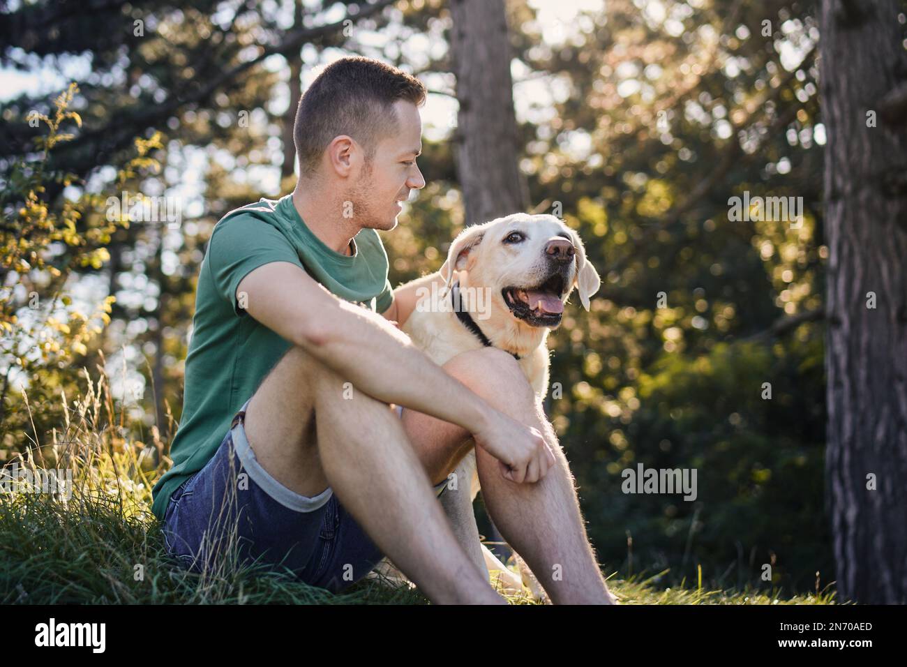 Uomo con il suo cane felice seduto in erba sotto l'albero. Il proprietario di un animale domestico che si diverte a fare un viaggio con il suo labrador Retriever durante la giornata estiva di sole. Foto Stock