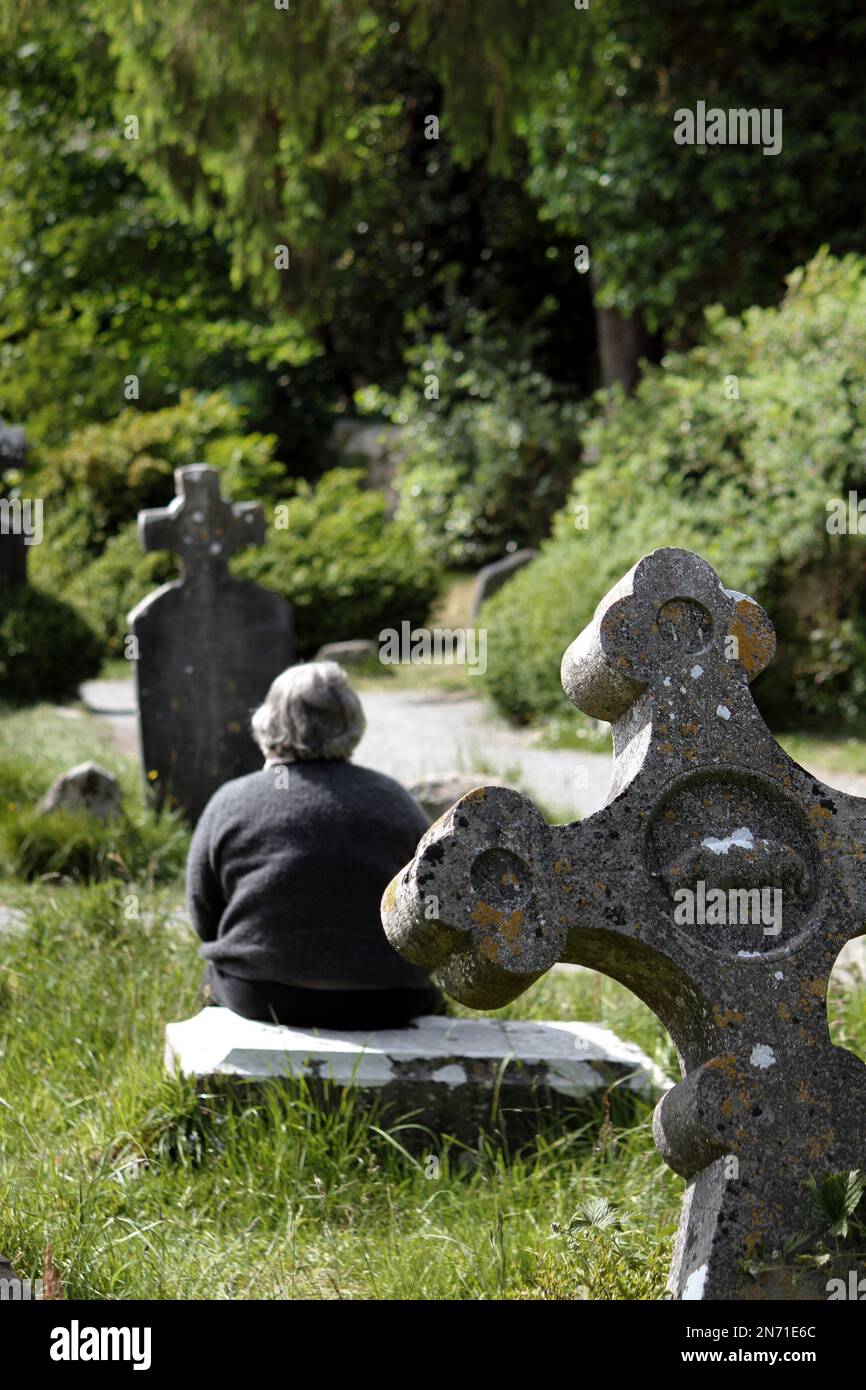 Donna seduta vicino alla croce di pietra medievale del monastero di Glendalough nelle montagne di Wicklow in Irlanda Foto Stock