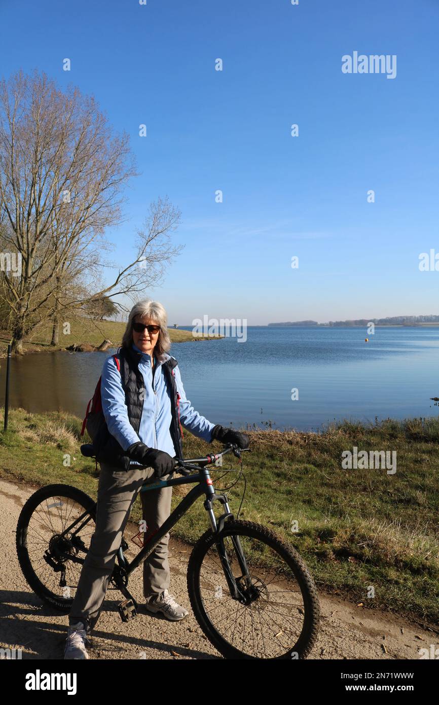 Donna degli anni sessanta che indossa occhiali da sole e guanti in bicicletta, ma senza aiuto intorno all'acqua di Rutland in inverno Foto Stock