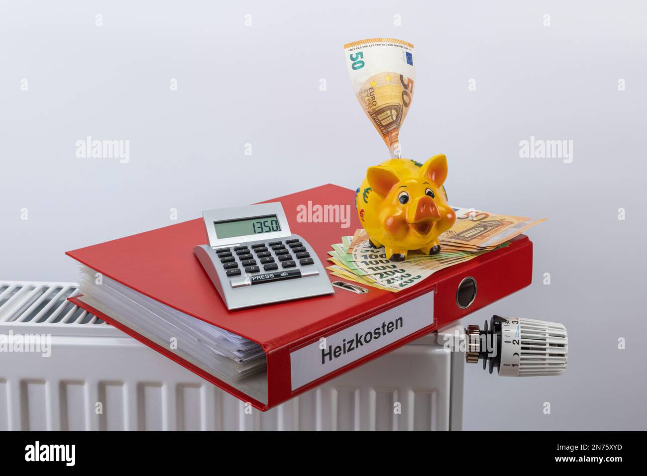 Cartella file rosso con etichetta costi energetici, calcolatrice tascabile, banca piggy, varie fatture in euro giacenti sul radiatore, 1350 euro, immagine simbolo, bolletta di riscaldamento, costi energetici, aumento dei costi di riscaldamento, sfondo bianco, Foto Stock