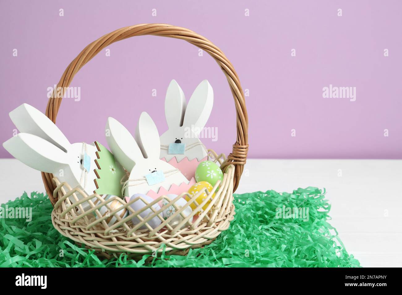 Coniglietti di legno con mascherine protettive, uova dipinte in cestello e erba di carta su tavolo di legno bianco. Vacanze di Pasqua durante la quarantena COVID-19 Foto Stock