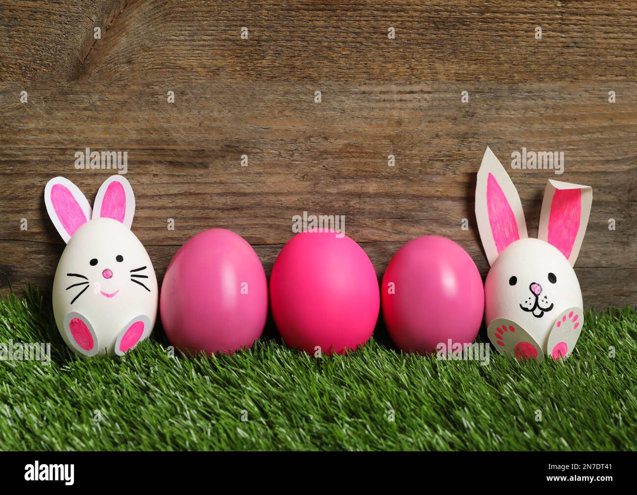 Due uova bianche come coniglietti pasquali tra gli altri su erba verde su sfondo di legno Foto Stock