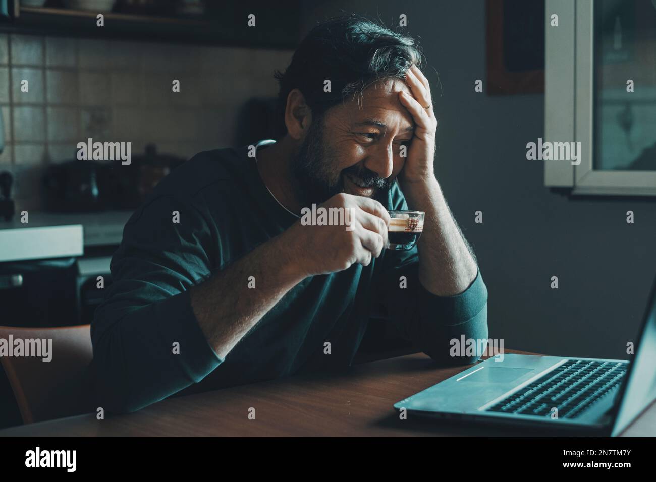 L'uomo da solo a casa usa un computer portatile sorridente e beve caffè espresso in cucina. Mattina presto lavoro e tecnologia internet indoor attività di svago c Foto Stock