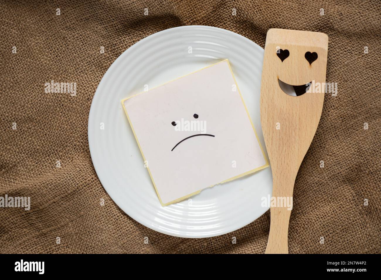 triste smiley giace su un foglio di carta su un piatto accanto ad una spatola di legno con un sorriso su uno sfondo marrone burlap, niente da mangiare, crisi Foto Stock
