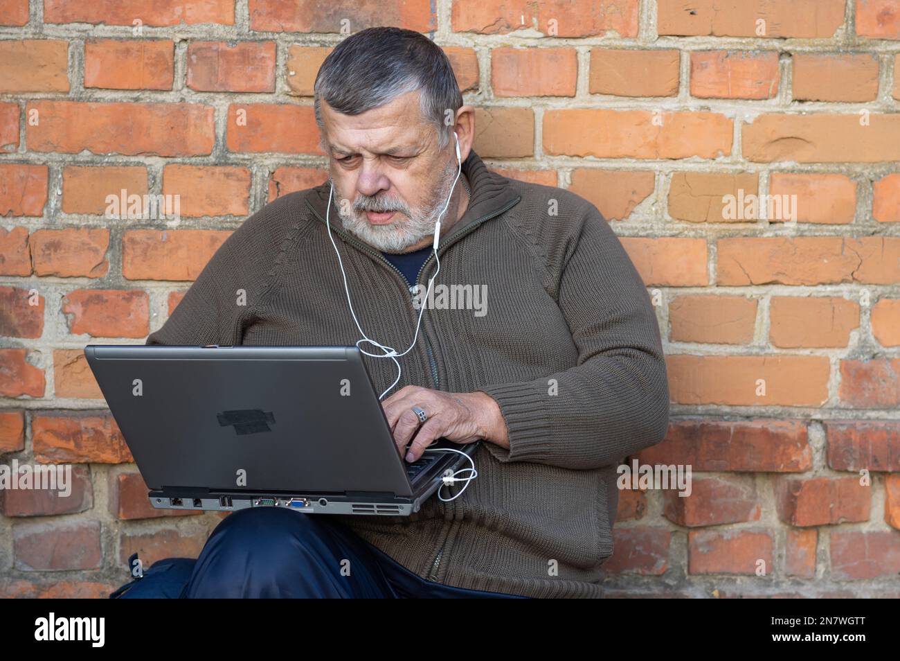 Bel ritratto del barbuto senior uomo al lavoro su un computer portatile e di ascoltare musica mentre seduto contro un muro di mattoni Foto Stock