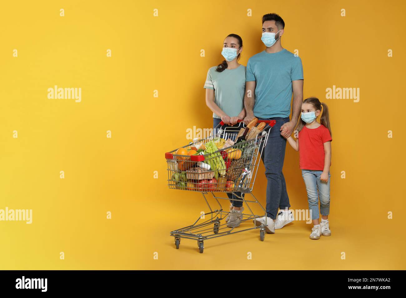 Famiglia con maschere protettive e carrello pieno di generi alimentari su sfondo giallo. Spazio per il testo Foto Stock