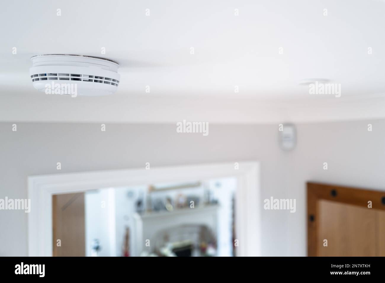 Messa a fuoco superficiale di un rivelatore di fumo cablato visto nella sala al piano inferiore, fissato al soffitto. Si può vedere una porta aperta che conduce ad un soggiorno. Foto Stock
