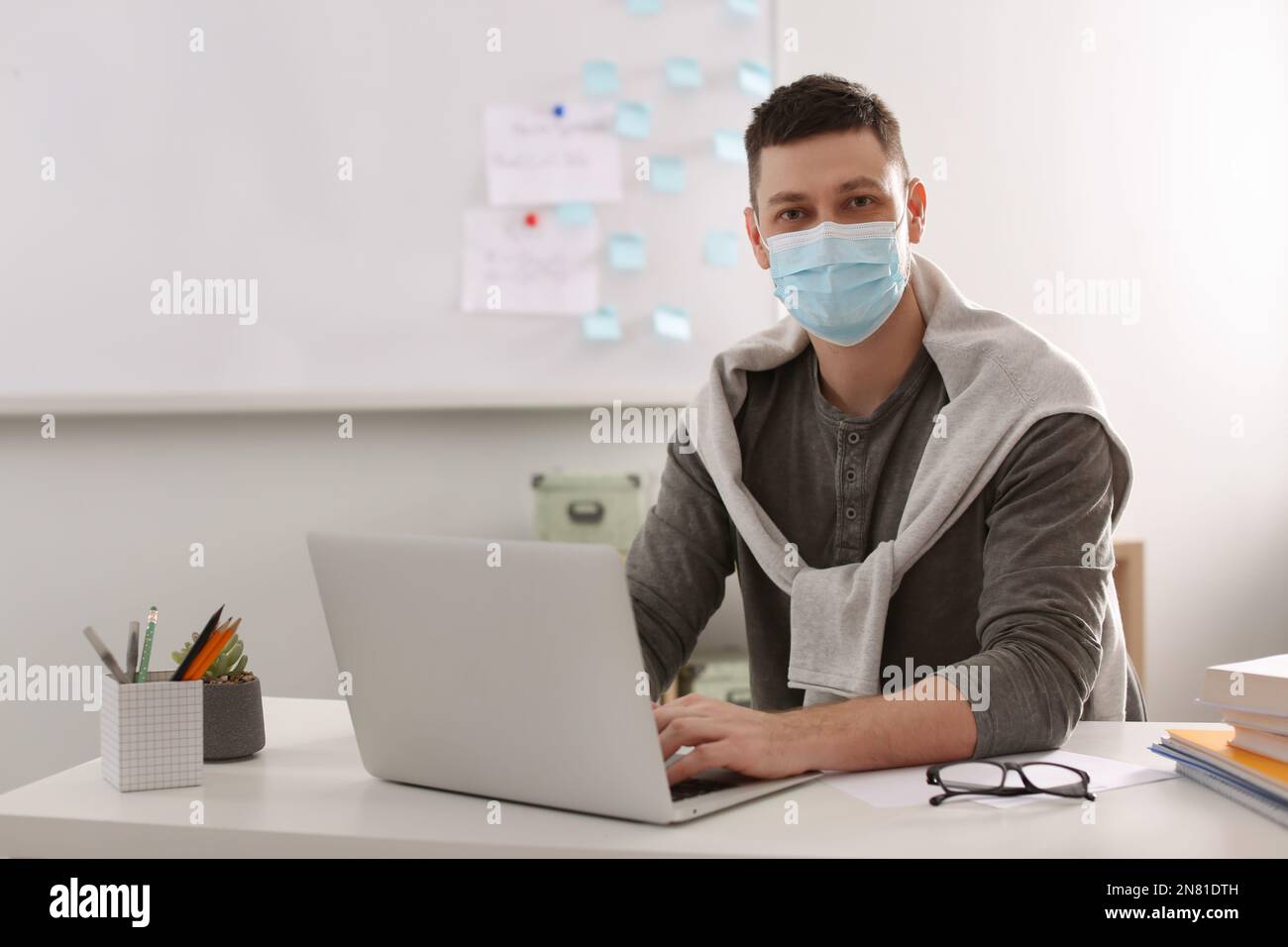 Insegnante con maschera protettiva e computer portatile seduto alla scrivania in aula. Riapertura dopo la quarantena di Covid-19 Foto Stock