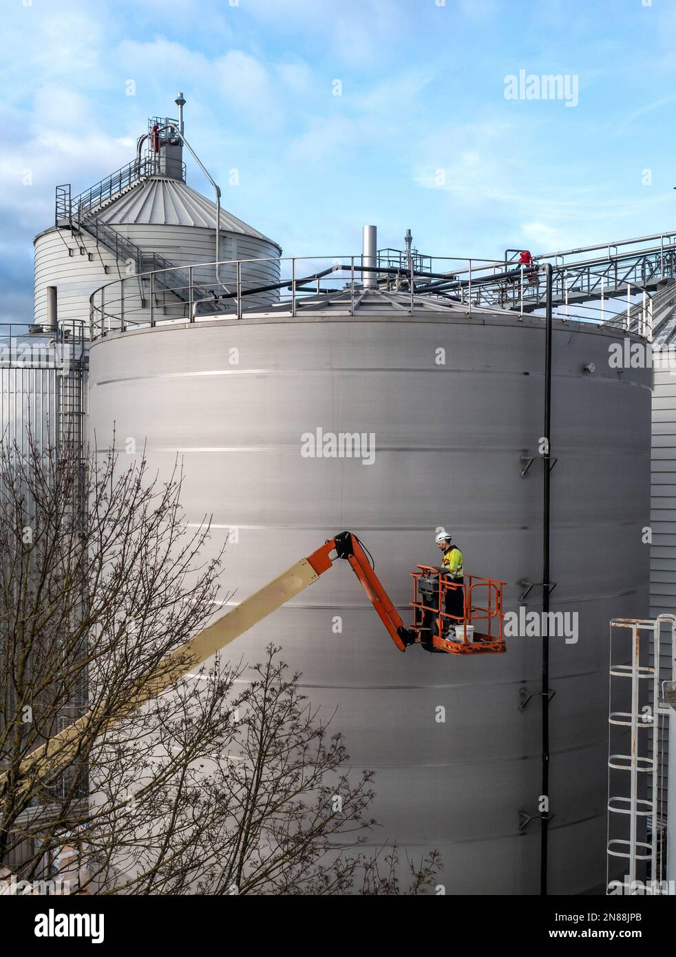 Un lavoratore maschio su una piattaforma di lavoro elevata o un raccoglitrice di ciliegie che lavora in altezza su grandi silos industriali o serbatoi di stoccaggio con spazio per le copie Foto Stock