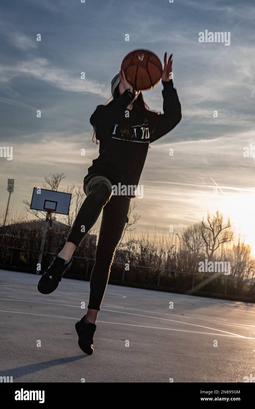 Vista frontale di una ragazza adolescente scatta una foto su un campo da basket, tramonto retroilluminato. Fotografia che cattura il sollevamento dei piedi da terra. Foto Stock