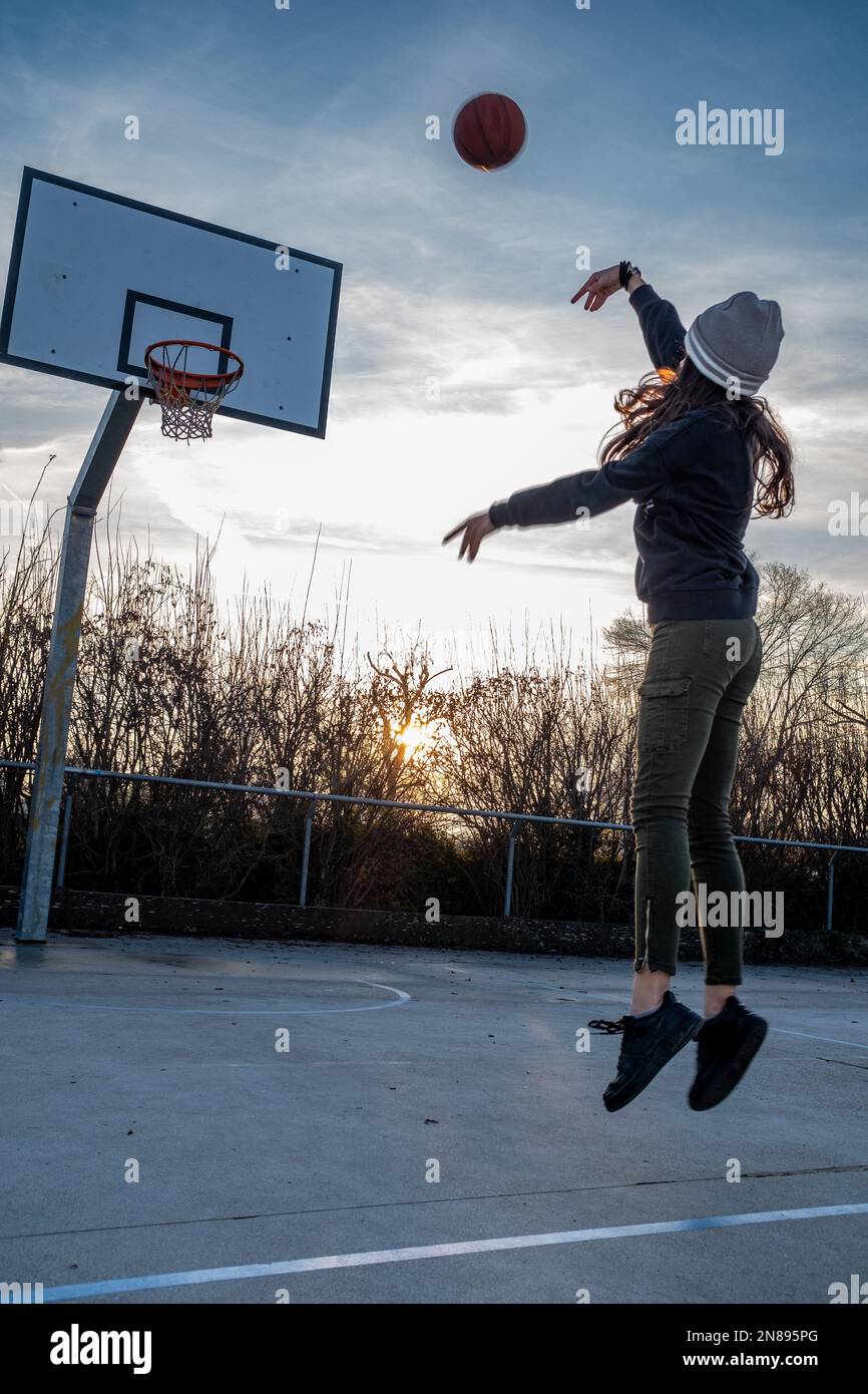 Vista posteriore di una ragazza adolescente, a mezz'aria, scatta una foto su un campo da basket, tramonto retroilluminato, composizione verticale, spazio copia. Foto Stock