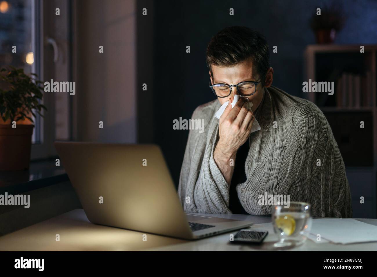 Uomo d'affari malato che soffia il naso mentre lavora al computer in ufficio a casa di notte. Il giovane uomo starnutisce con il naso che corre mentre lavora da casa in auto-isolamento, ha freddo cattivo, influenza, febbre. Rimani a casa Foto Stock