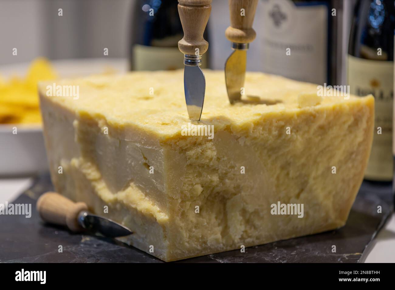 Primo piano del tipico Parmigiano Reggiano italiano con coltello tipico per staccare i pezzi , sullo sfondo bottiglie di vino sfocate Foto Stock