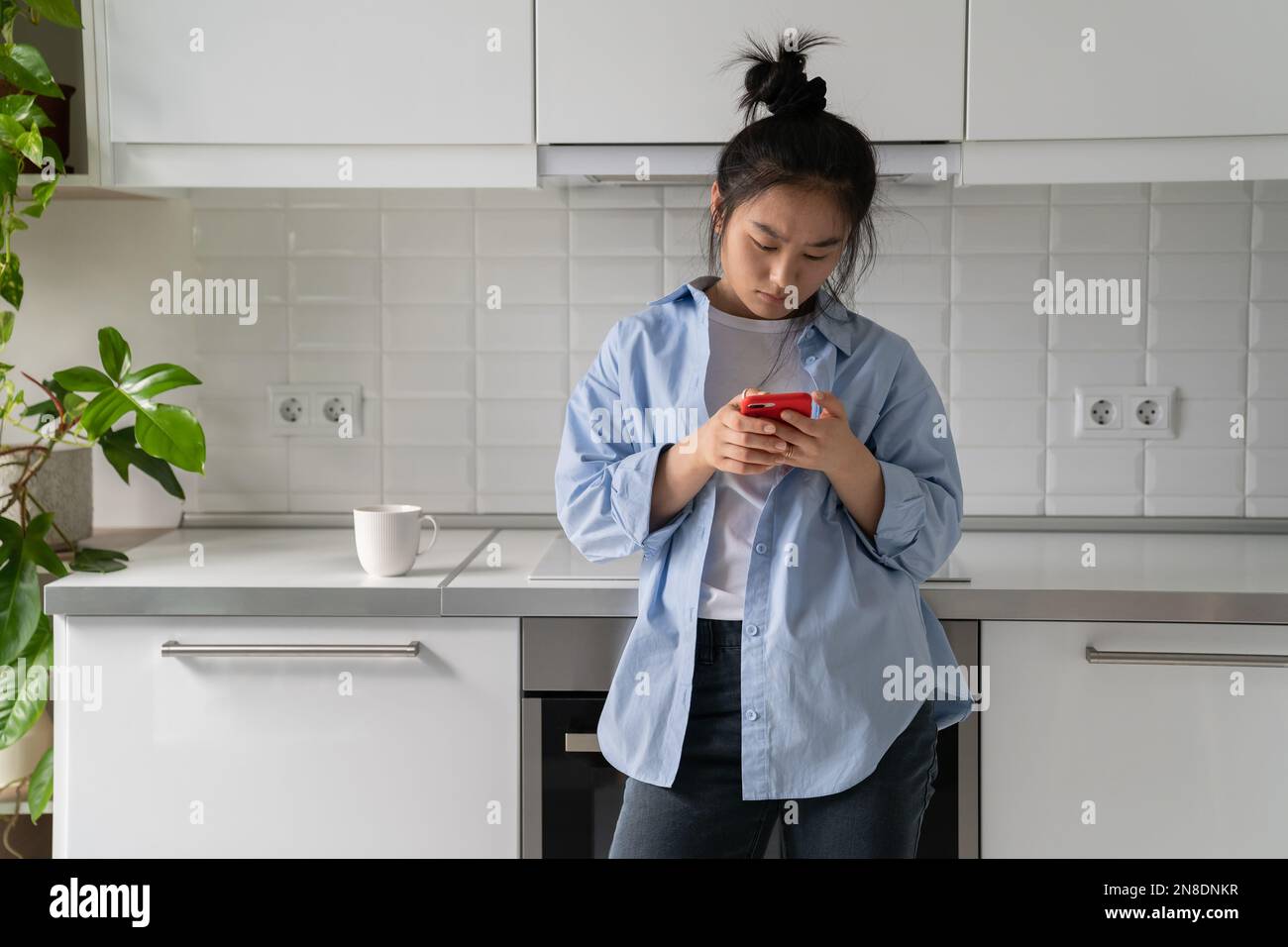 Donna asiatica concentrata emotionless digitando testo sul telefono in piedi in cucina con caffè di vetro Foto Stock