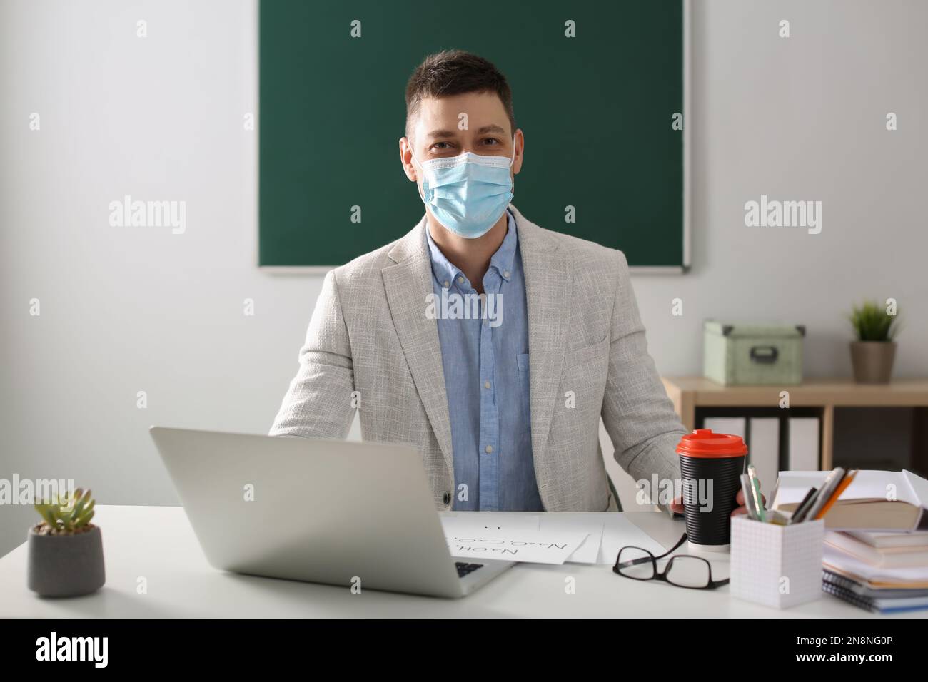 Insegnante con maschera protettiva e computer portatile seduto alla scrivania in aula. Riapertura dopo la quarantena di Covid-19 Foto Stock