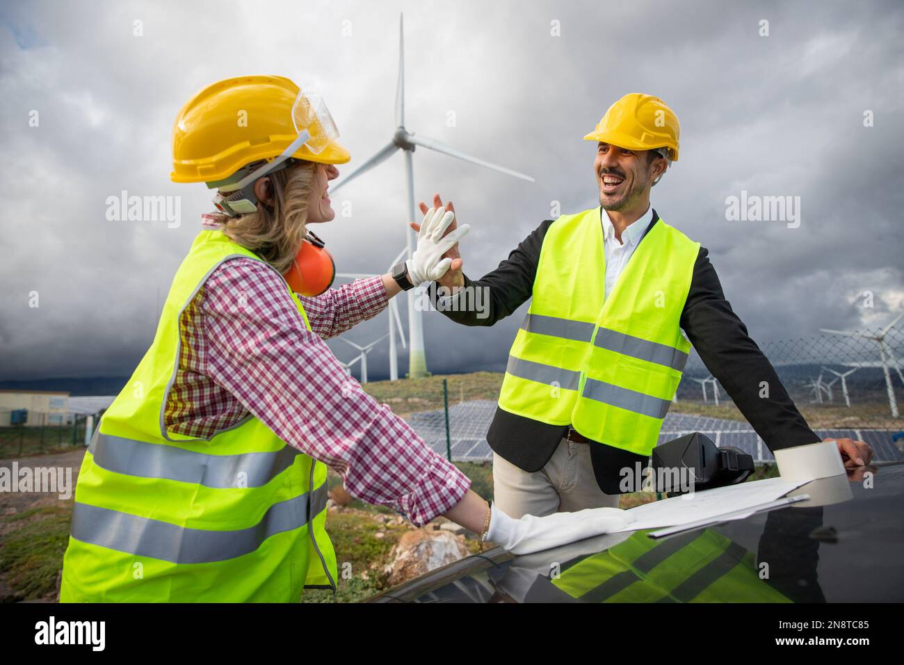 Due colleghi soddisfatti si danno a vicenda un alto cinque mentre lavorano consultando un disegno tecnico in una fattoria solare. Foto Stock Due colleghi soddisfatti si danno a vicenda un alto cinque mentre lavorano consultando un disegno tecnico in una fattoria solare. Foto Stock