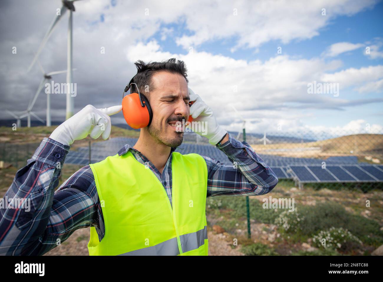Un lavoratore soffre di dolore all'orecchio vicino alle turbine eoliche, quindi indossa cuffie a cancellazione del rumore. Foto Stock Un lavoratore soffre di dolore all'orecchio vicino alle turbine eoliche, quindi indossa cuffie a cancellazione del rumore. Foto Stock