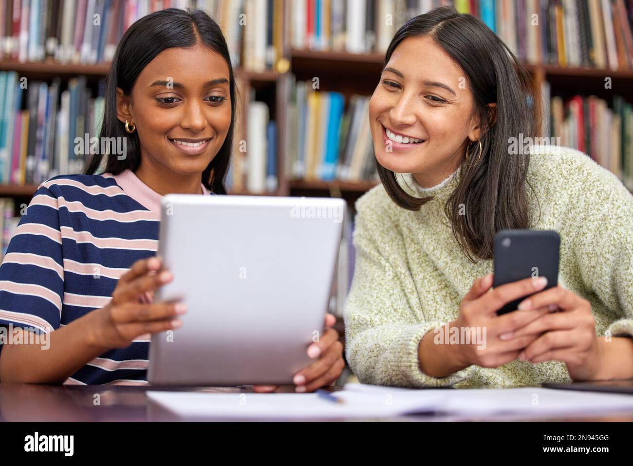 Trova la tua cosa. due femmine che usano i loro dispositivi in una biblioteca. Foto Stock
