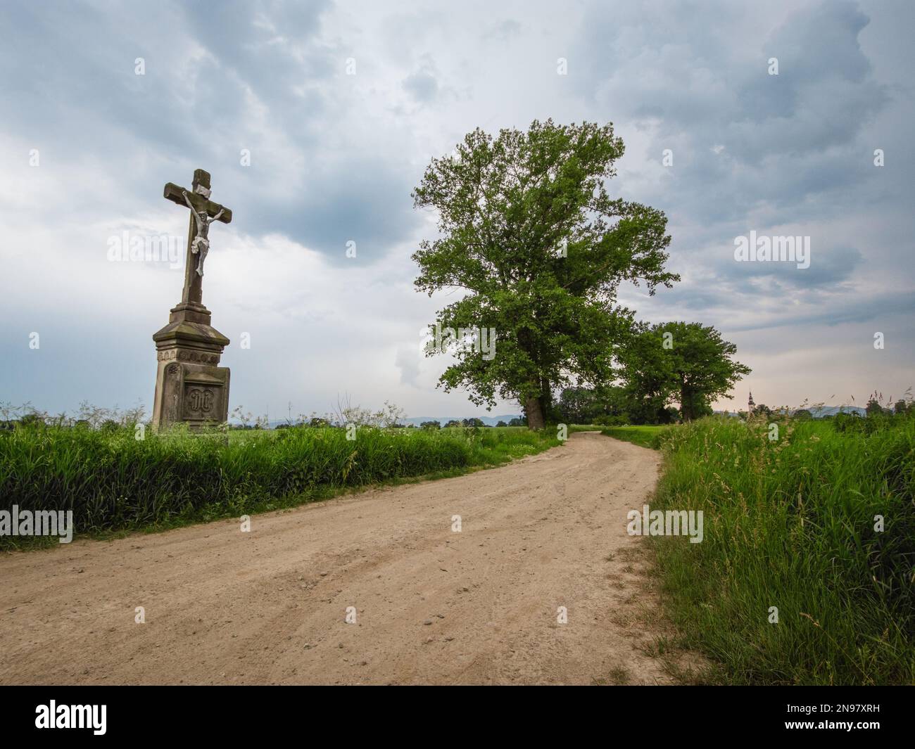 Campo, strada sterrata con una croce a bordo strada. Estate, paesaggio rurale con croce di pietra, prato e alberi. Tempo nuvoloso, senza corpo, Polonia. Foto Stock