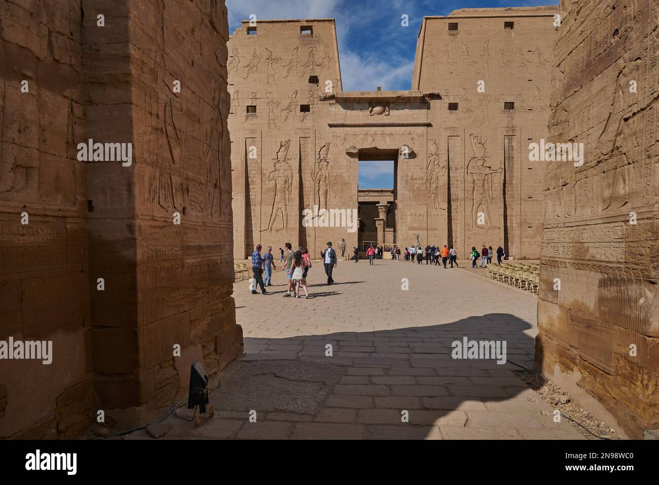 Il Tempio di Edfu a Edfu, Egitto, vista esterna della luce del giorno che mostra l'ingresso principale con i visitatori e le nuvole nel cielo sullo sfondo Foto Stock