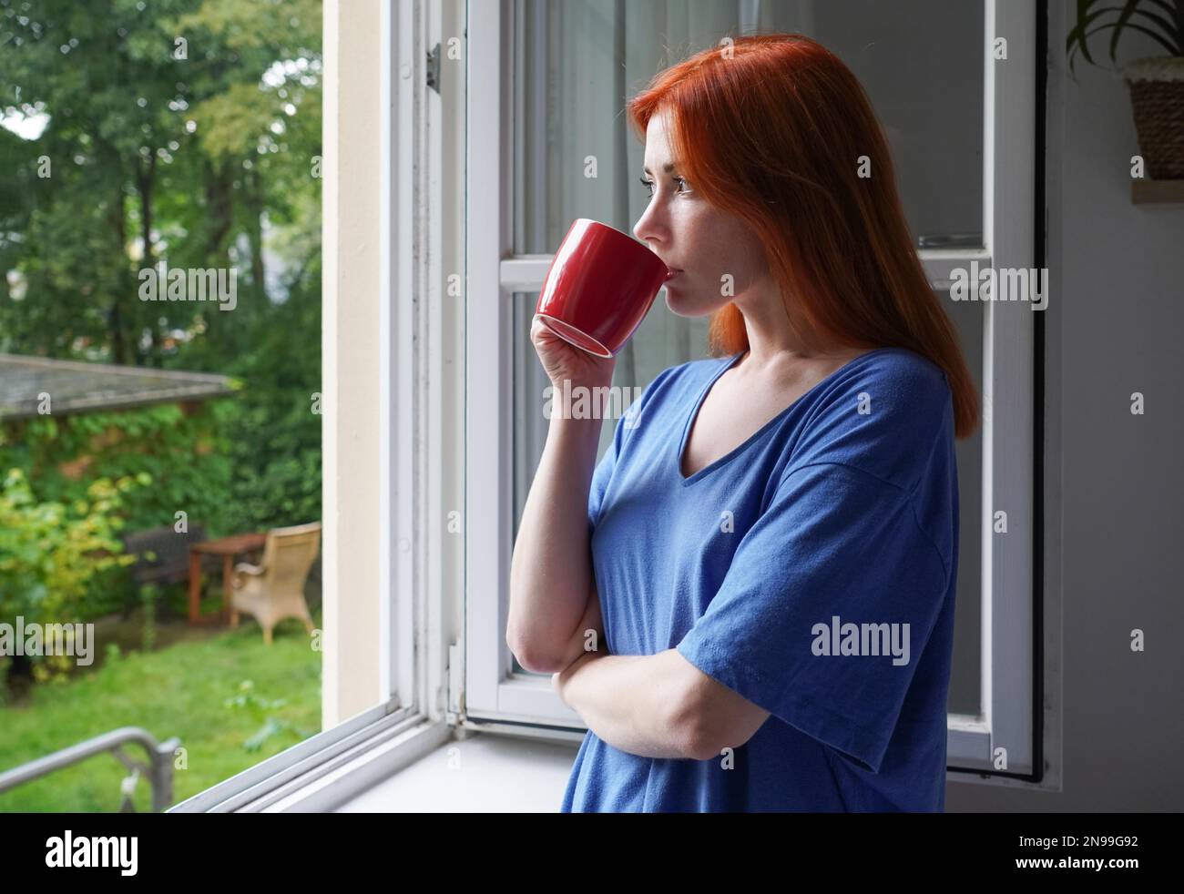 giovane donna in piedi dalla finestra aperta bevendo dalla tazza di caffè mentre guarda fuori nel giardino Foto Stock