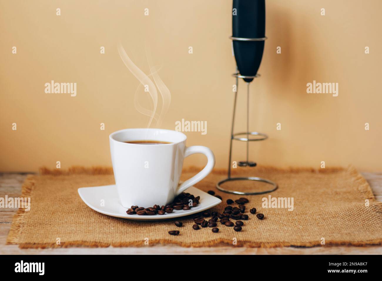 Tazza bianca di caffè caldo con chicchi su tovagliolo da sacco e pannarello su tavolo di legno contro parete beige. Messa a fuoco selettiva. Foto Stock