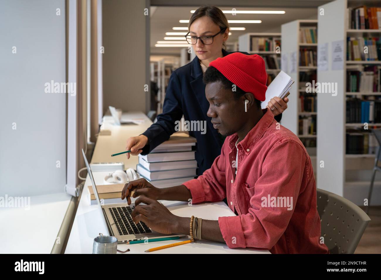 Insegnante donna che indica lo schermo del notebook mentre spiega qualcosa allo studente africano in biblioteca Foto Stock