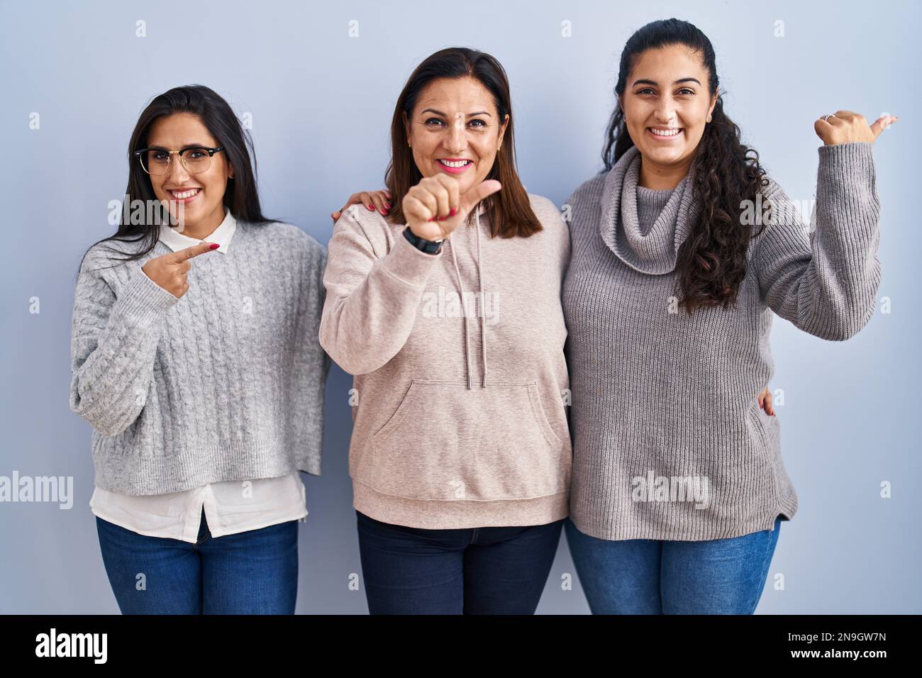 Madre e due figlie in piedi su sfondo blu puntando alla schiena dietro con la mano e pollici in su, sorridendo fiducioso Foto Stock