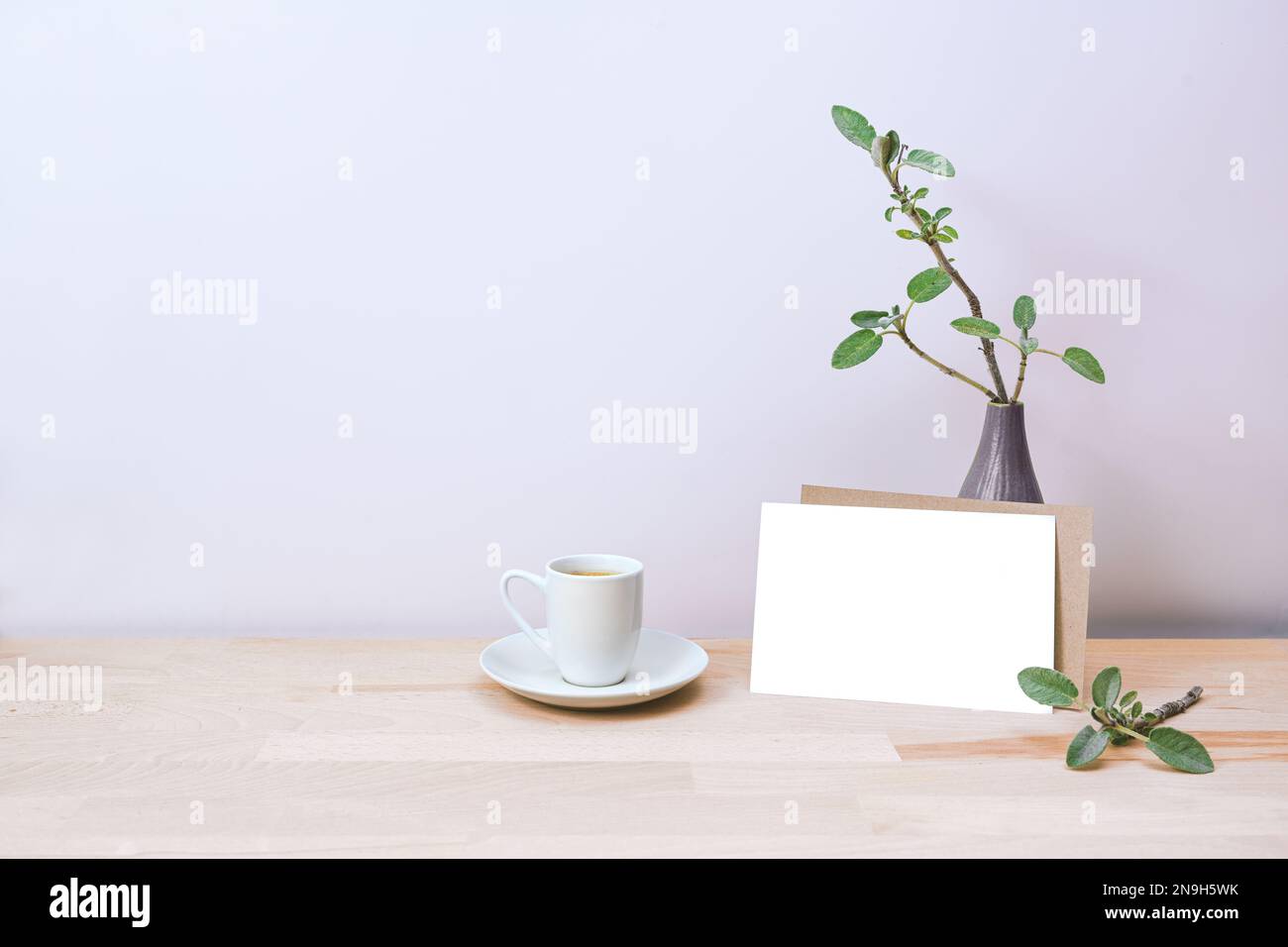 Scheda vuota per l'invito o la mock-up di saluto appoggiata su un vaso con pochi rami e una tazza di caffè, su un tavolo di legno contro un pallido muro di lavanda, mi Foto Stock