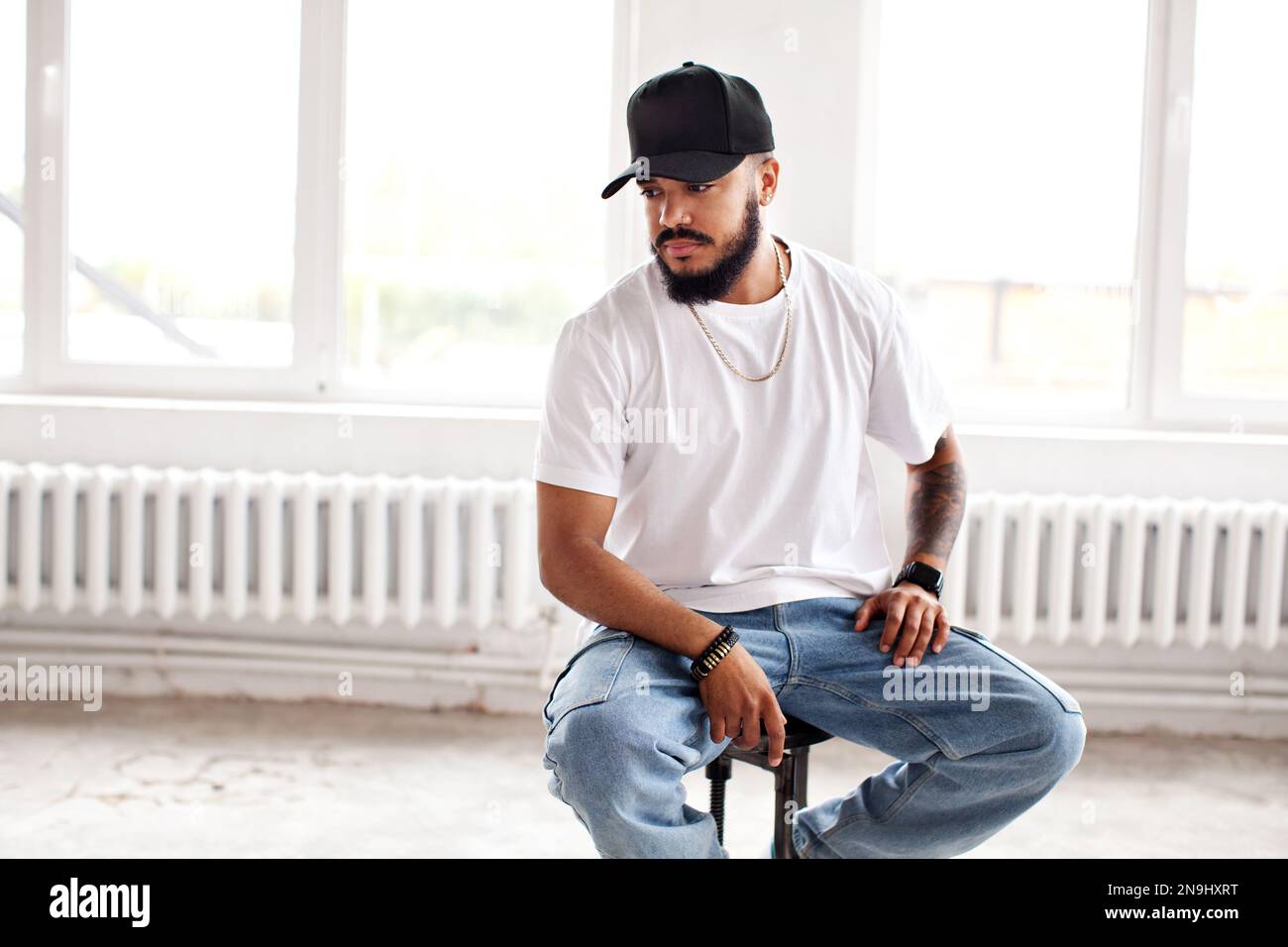 Autentico uomo portatore seduto su sfondo bianco loft, guardando lontano, indossando casual T-shirt bianca, berretto da baseball e jeans, stile di vita Foto Stock