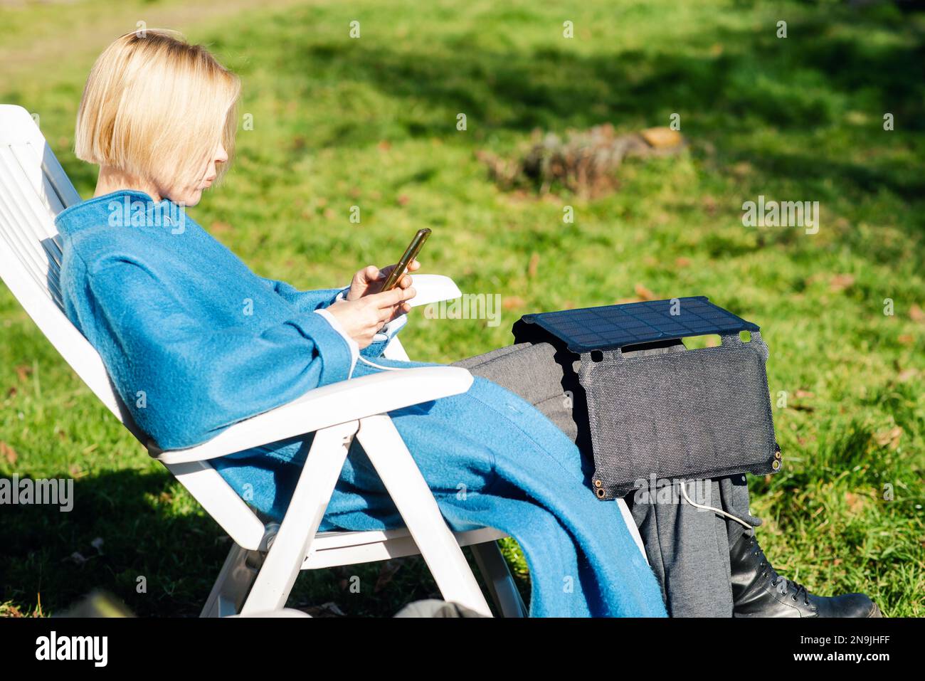 Una donna in un cappotto con un telefono cellulare in mano e una batteria solare portatile siede all'aperto nel giardino. Concetto ecologico di energia alternativa. Foto Stock